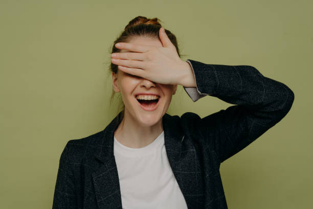 Laughing young female in casual clothes covering eyes with hand while waiting for surprise Laughing young female in casual clothes covering eyes with hand while waiting for surprise, smiling teenage girl keeping mouth opened with embarrassed expression, studio shot hair covering eyes stock pictures, royalty-free photos & images