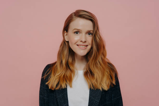 Positive young woman with ginger hair smiling at camera posing against pink wall Photo of pretty smiling female with ginger hair without makeup looking at camera wearing dark formal jacket and white top, happy teenage girl posing isolated over pink studio background medium length hair stock pictures, royalty-free photos & images