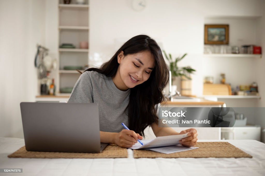 Beautiful Mongolian woman taking care of her finances at home Young woman of a Mongolian ethnicity, doing her taxes and sorting out her finances at home, using a laptop Home Finances Stock Photo