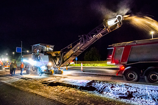 Road reconstruction works at night. Cold milling machine removing asphalt pavement and loading it into the dump tuck. The works are taking place on a partially closed highway. Traffic is slow, but passing vehicles can be seen in the background.