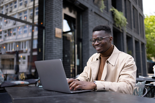 Handsome African American male entrepreneur relaxing while sitting in a coffee shop, casually working using a laptop and enjoying himself and smiling