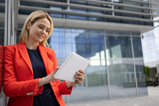 Happy middle aged Caucasian businesswoman casually using a digital tablet computer outdoors, smiling and standing in front of her office building