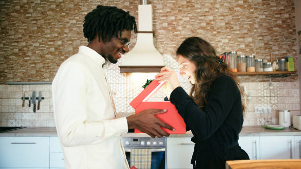 istock Cheerful man surprising his girlfriend with gift at home in the kitchen 1324749621