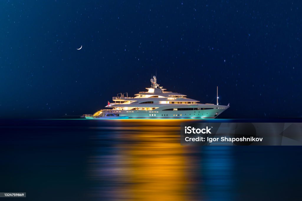 Night view to large illuminated white boat located over horizon, colorful lights coming from yacht reflect on the surface of the the Gulf sea. Shot at blue hour. Yacht Stock Photo