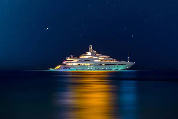 Photo of Night view to large illuminated white boat located over horizon, colorful lights coming from yacht reflect on the surface of the the Gulf sea. Shot at blue hour.