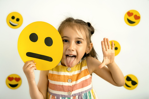 A funny girl holds a cardboard sad emoticon in her hands and shows her tongue while laughing. World emoji day. Anthropomorphic smile Face.