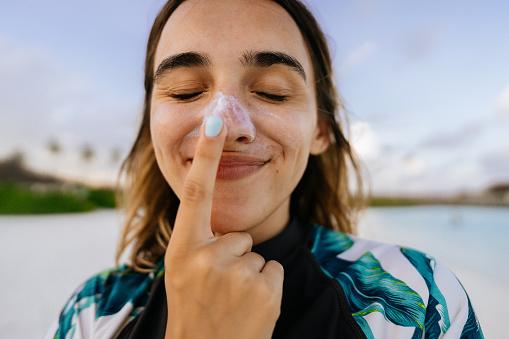 Photo of a young woman applying sunscreen while being outdoors, at the beach; making silly faces while reapplying SPF and protecting her skin from the sun.