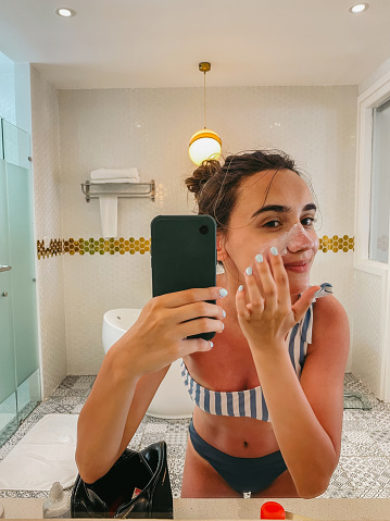 Photo of a young woman applying sunscreen and taking mirror selfies in the bathroom; making silly faces while applying SPF and protecting her skin from the sun before going out; mobile stock photo made with iPhone 12 Pro.