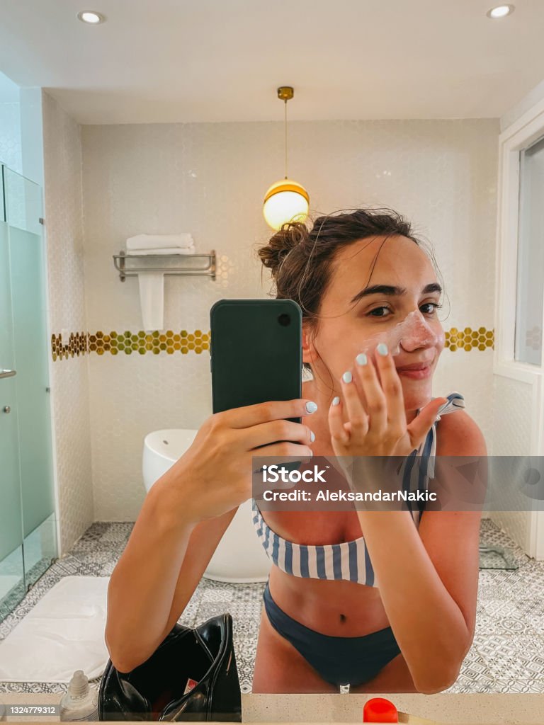 Wearing my SPF! Photo of a young woman applying sunscreen and taking mirror selfies in the bathroom; making silly faces while applying SPF and protecting her skin from the sun before going out; mobile stock photo made with iPhone 12 Pro. Selfie Stock Photo