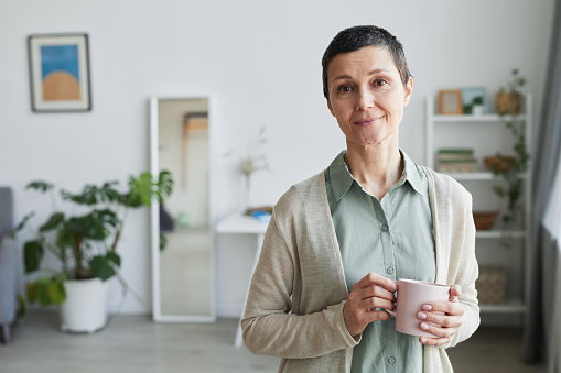 Waist up portrait of mature woman smiling at camera while standing by window and holding cup of coffee at home, copy space