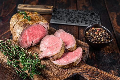 Roast Tenderloin beef fillet meat on a wooden board with herbs. Dark wooden background. Top view.