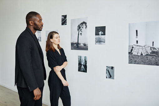 Portrait of elegant mixed-race couple wearing black while looking at photographs in modern art gallery, copy space