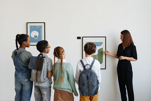 Back view at diverse group of children listening to female tour guide while visiting modern art gallery, copy space