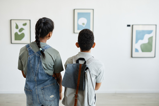 Back view at two schoolchildren visiting art gallery and looking at modern abstract paintings, copy space