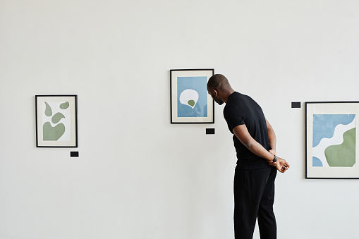 Minimal back view at African-American man looking at paintings in modern art gallery, copy space