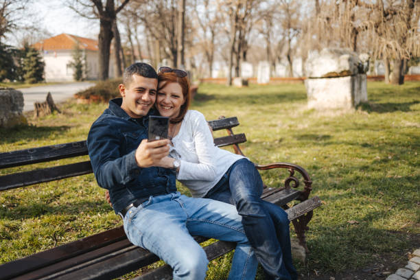 счастливая молодая влюбленная пара сидит на скамейке в общественном парке и делает селфи на мобильном телефоне - desire men young women tree стоковые фото и изображения