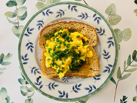 High angle closeup view of golden scrambled organic eggs with fresh chives on a slice of buttered, toasted sourdough bread on a round, leaf pattern ceramic plate