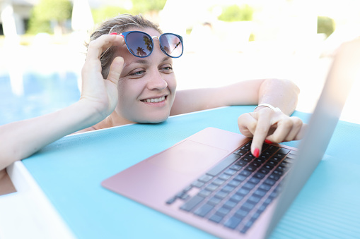 Young smiling woman is working on laptop while in pool. Remote work and telework concept