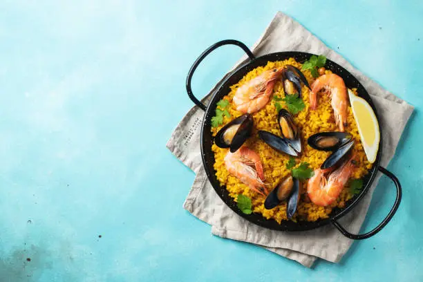 Traditional spanish seafood paella in pan with chickpeas, shrimps, mussels, squid on light blue concrete background. Top view with copy space.