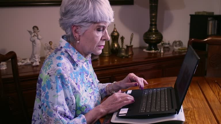 A Senior Latin American Lady Works From Home On Her Laptop Computer Sitting At Her Dining Table