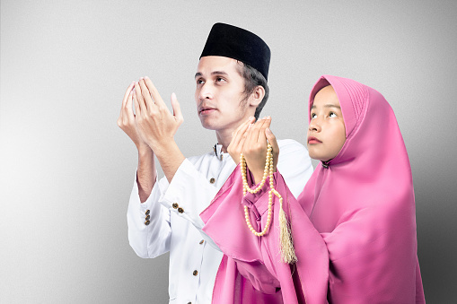 Asian Muslim couple standing while raised hands and praying together with gray wall background