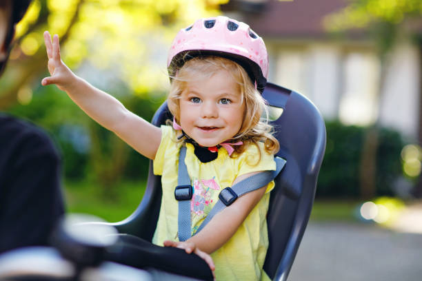portrait of little toddler girl with security helmet on the head sitting in bike seat of parents. boy on bicycle on background. safe and child protection concept. family and weekend activity trip. - helmet bicycle little girls child imagens e fotografias de stock