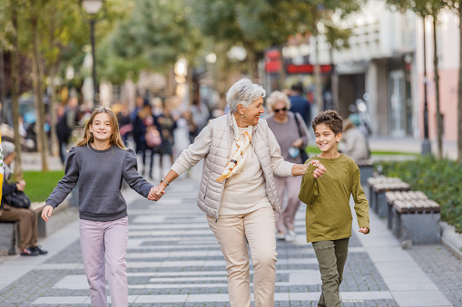 An Older Woman is Enjoying the Time with her Grandson and Granddaughter Holding Hands and Walking Down the City District.