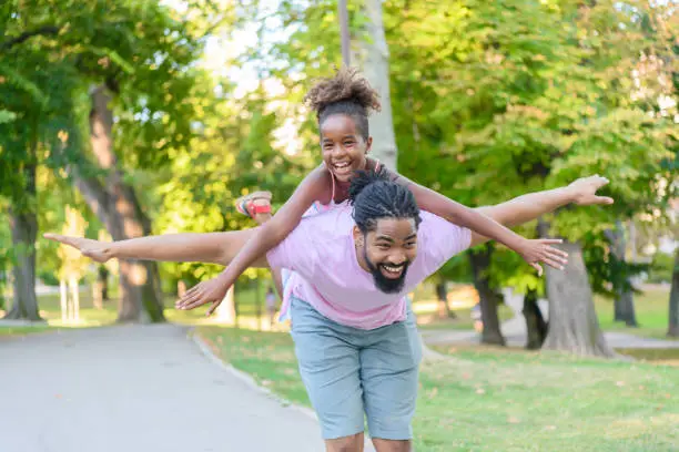 Photo of Cheerful African-American Father is Carrying his Daughter on Shoulders in Public Park and Playing.