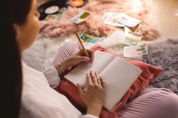 High angle view of young woman sitting comfortably on bedroom floor and writing memories in her diary.