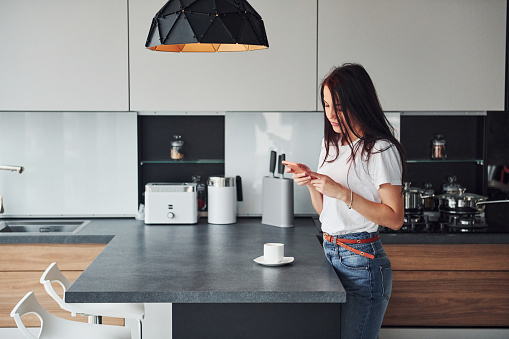 Young beautiful brunette in casual clothes indoors in kitchen at daytime using smartphone.