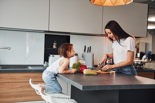 Mother with her little daughter slicing vegetables indoors in kitchen.