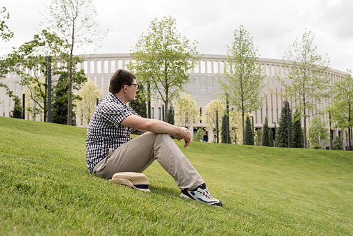 Young thoughtful man in summer hat sitting on the grass in the park, looking away