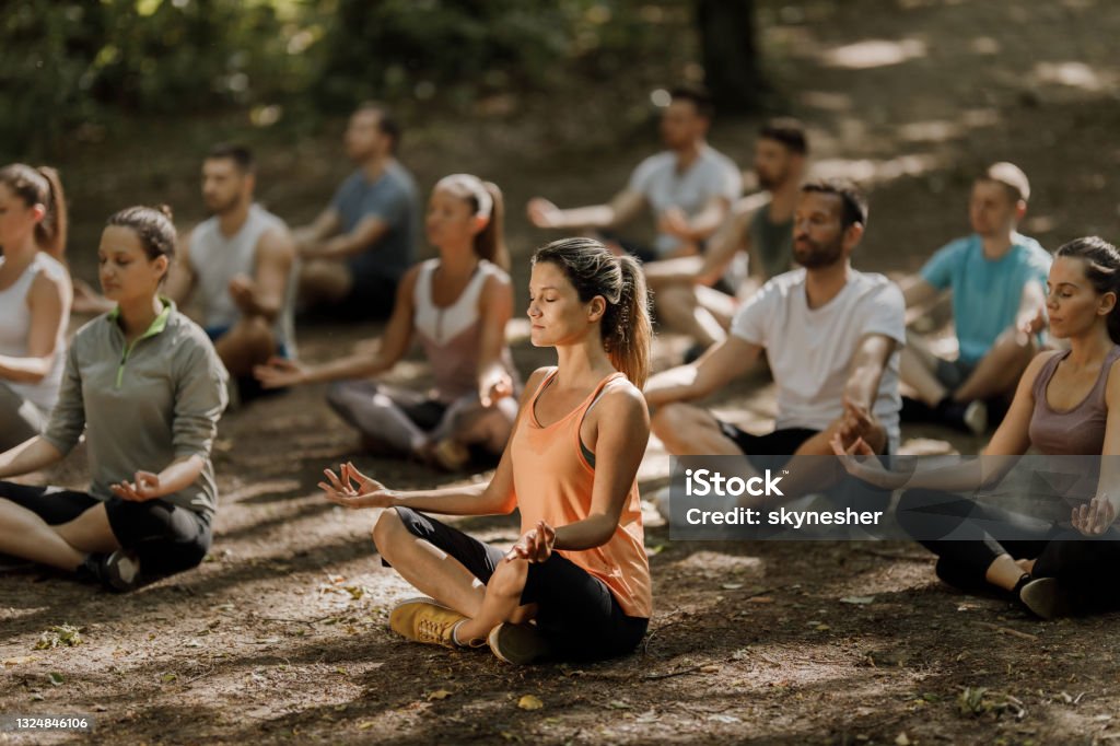 Large group of athletes meditating on Yoga class in nature. Group of Zen-like people sitting in Lotus position while exercising Yoga with their eyes closed in the park. Focus is on woman in the foreground. Meditating Stock Photo