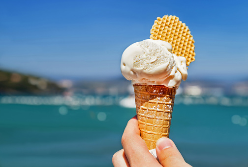 Delicious ice cream cone held up on the blurry sea and sky background. Tasty, sweet, fresh food for people in the summer. Copy space.