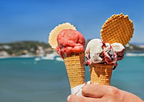Two delicious ice cream cones held up on the blurry sea and sky background. Tasty, sweet, fresh food for people in the summer. Copy space.