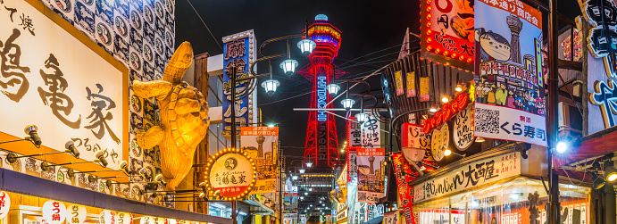 The iconic spire of the Tsutenkaku Tower overlooking the colourful neon lights illuminating the bars and restaurants of Shinsekai at night in the heart of Osaka, Japan’s vibrant second city.