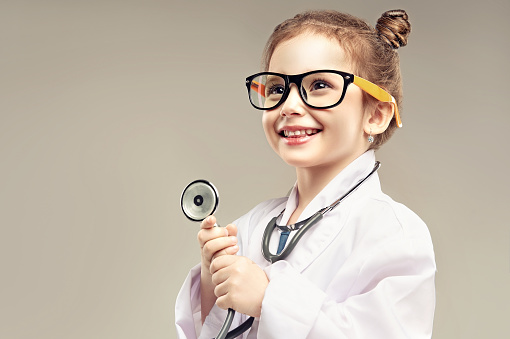 Small preschool girl is playing a doctor. Toothy smiling girl dressed in medicine white coat and big eyeglasses is holding stethoscope in the hands. Role playing games. Happy childhood.