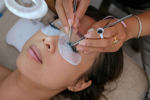 Close-up shot of a beautiful Asian woman getting home service eyelashes extension. She is closing her eyes while an unrecognizable beautician carefully putting on the extension with tweezers.