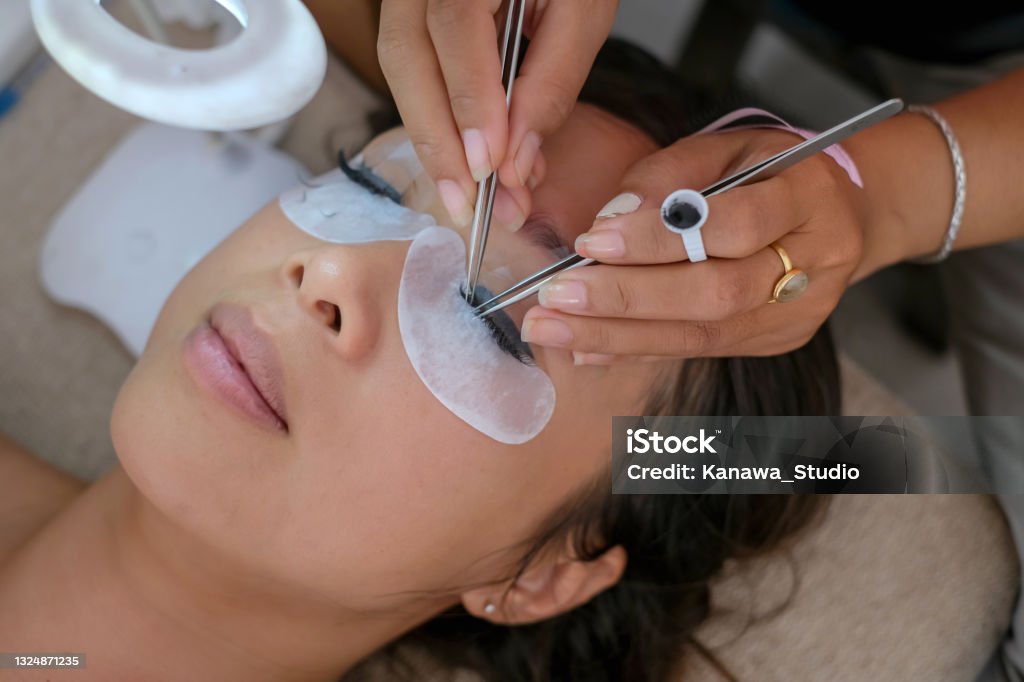 Beautiful Woman Getting Home Service Eyelash Extension Close-up shot of a beautiful Asian woman getting home service eyelashes extension. She is closing her eyes while an unrecognizable beautician carefully putting on the extension with tweezers. False Eyelash Stock Photo