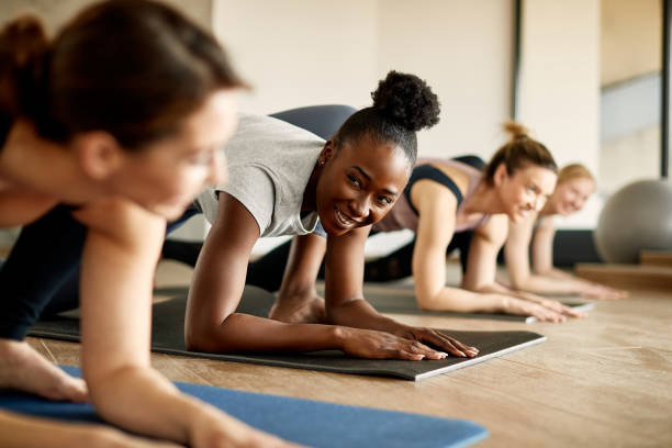 Happy African American woman talking to her friend during exercise class at health club. Group of athletic woman warming up on he floor at health club. Focus is on African American woman. exercise class stock pictures, royalty-free photos & images
