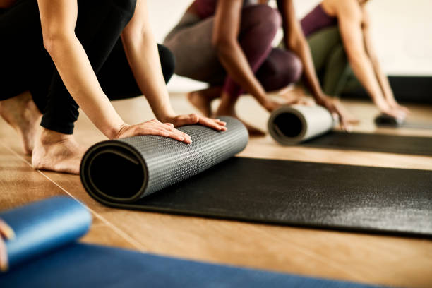 Close-up of athletic woman rolling up her exercise mat after practicing at health club. Close-up of athletic women preparing their exercise mats for sports training at health club. Focus is on woman in black. mat stock pictures, royalty-free photos & images