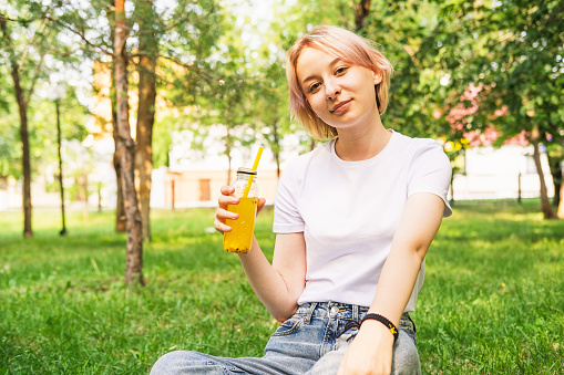The girl drinks orange juice on a background of juicy greenery. Beautiful teenage girl in the park. Stylish slender girl leads a healthy lifestyle. Fresh young face