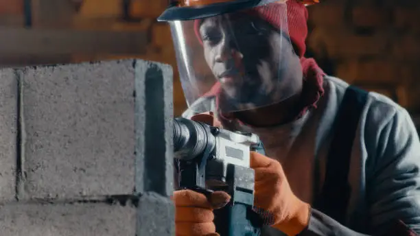 Black man in hardhat with face shield drilling hole in concrete brick while working on construction site