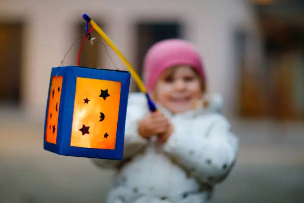Photo of Close-up of little kid girl holding selfmade lanterns with candle for St. Martin procession. Healthy toddler child happy about children and family parade in kindergarten. German tradition Martinsumzug