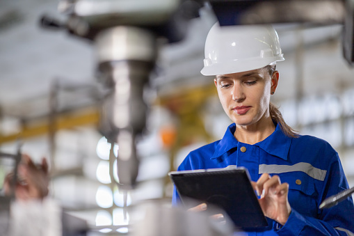 Caucasian White Female Engineer controls the quality using a tablet device to inspect the drilling machine in the factory.
