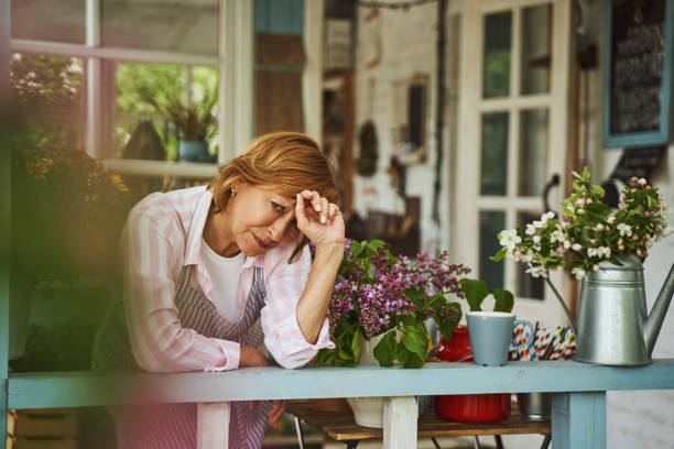 Upset aged female in apron is standing on veranda of flower shop in village among blossom