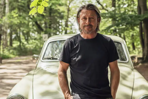 Photo of Man in a black t-shirt standing by a classic car on a forest dirt road.