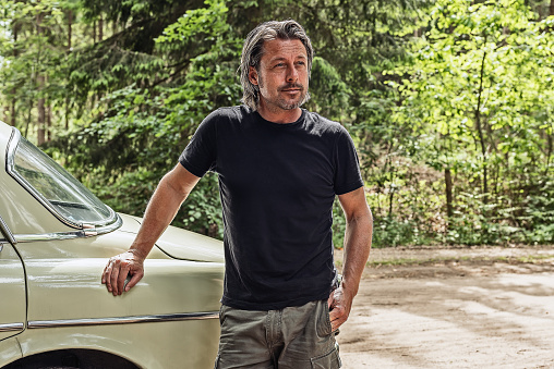 Man in a black t-shirt standing by a classic car on a forest dirt road.