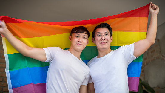 Portrait of young asian men couple showing their love with rainbow flag at home. LGBT, Gay and love relationship concept
