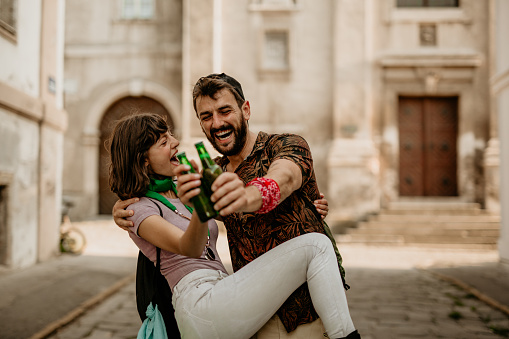 A spontaneous photo of fun and cheerful young adult brunette with bangs, wearing a purple top with colorful scarfs, singing and dancing down the street with her cool looking bearded friend, holding beers and smiling, wearing colorful shirt and a cap. Excitement grows in the new city walls